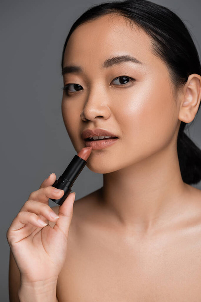 Portrait of young asian woman with naked shoulder holding lipstick isolated on grey  - Foto, Imagen