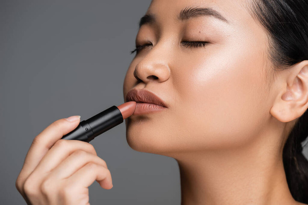 Young asian woman with makeup applying lipstick isolated on grey  - Foto, imagen