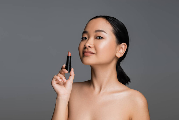 Pretty asian woman with naked shoulders holding beige lipstick isolated on grey  - Zdjęcie, obraz
