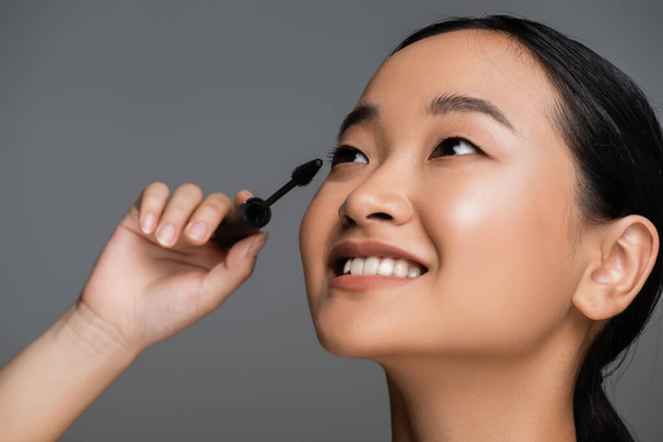 portrait of young asian woman with perfect smile applying black mascara isolated on grey - Photo, image