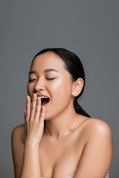 young asian woman with perfect skin and bare shoulders covering mouth with hand while yawning with closed eyes isolated on grey - Fotografie, Obrázek