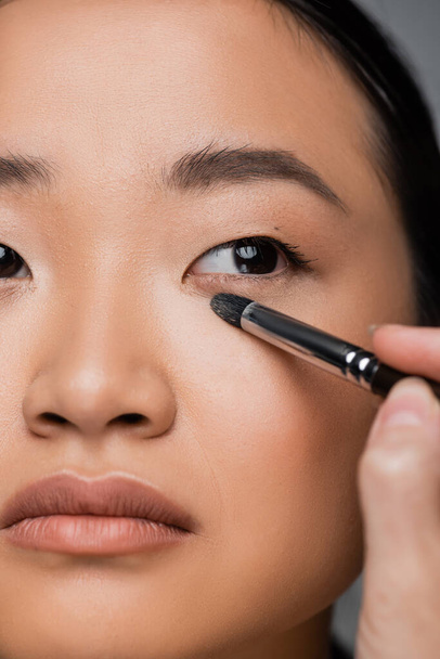 close up view of cropped asian woman with makeup foundation applying eye shadow isolated on grey - Photo, image