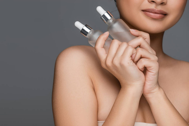partial view of woman with perfect skin holding bottles with cosmetic serum isolated on grey - Foto, immagini