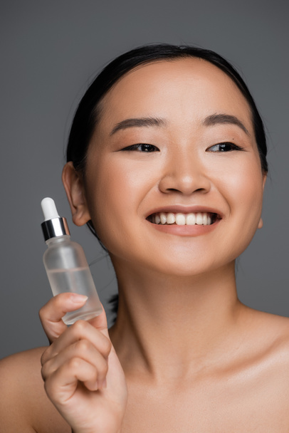 νεαρή και ευτυχισμένη Ασιάτισσα γυναίκα με καθαρό δέρμα που κρατά μπουκάλι καλλυντικού ορού που απομονώνονται σε γκρι - Φωτογραφία, εικόνα