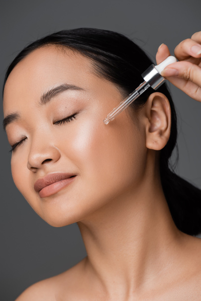 young asian woman with closed eyes applying cosmetic serum on clean face isolated on grey - Foto, imagen