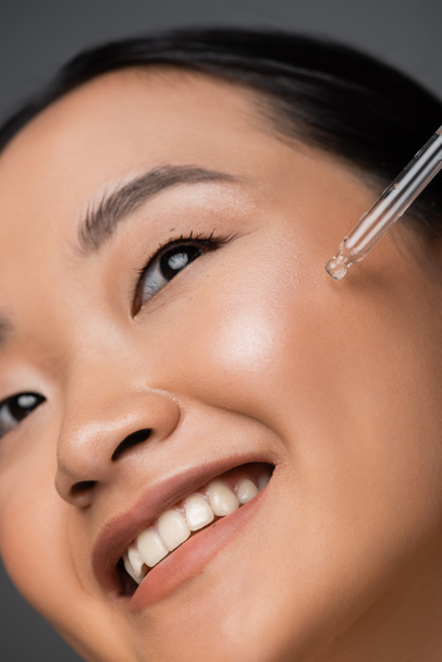 close up view of smiling asian woman applying cosmetic serum during facial pampering isolated on grey - Fotó, kép