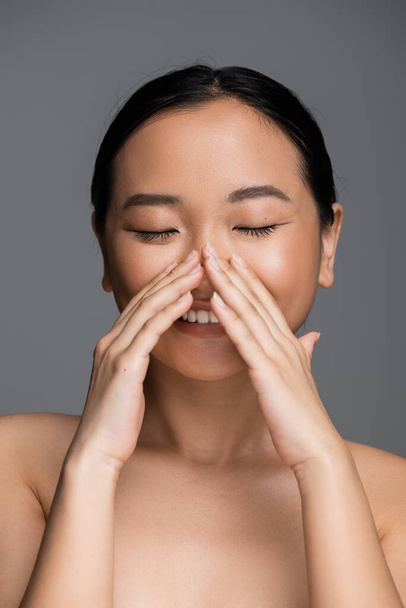 happy asian woman with closed eyes and perfect skin touching face isolated on grey - Fotoğraf, Görsel