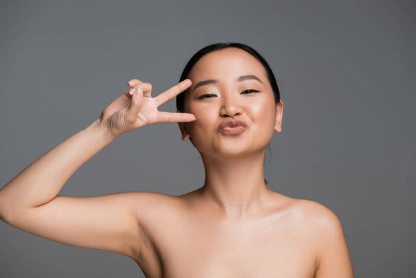 asian woman with bare shoulders pouting lips and showing victory gesture near face isolated on grey - Fotografie, Obrázek