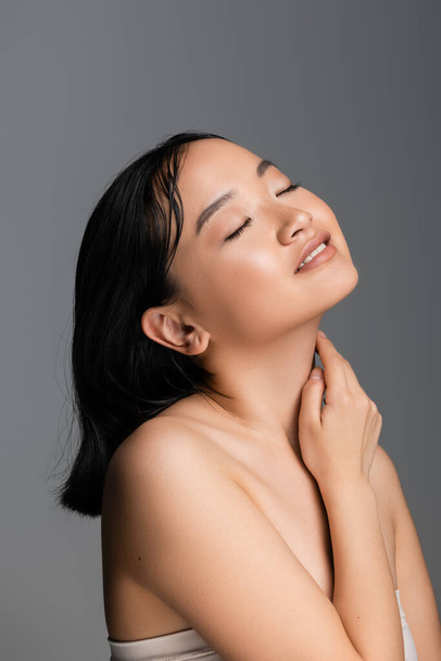 задоволена азіатська жінка з закритими очима та оголеним макіяжем, торкаючись шиї, ізольована на сірому
 - Фото, зображення
