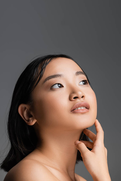 portrait of brunette asian woman with perfect skin and natural makeup touching neck and looking away isolated on grey - Foto, afbeelding