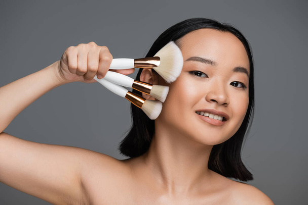 happy asian woman with bare shoulders and natural makeup holding different cosmetic brushes isolated on grey - Foto, Imagem