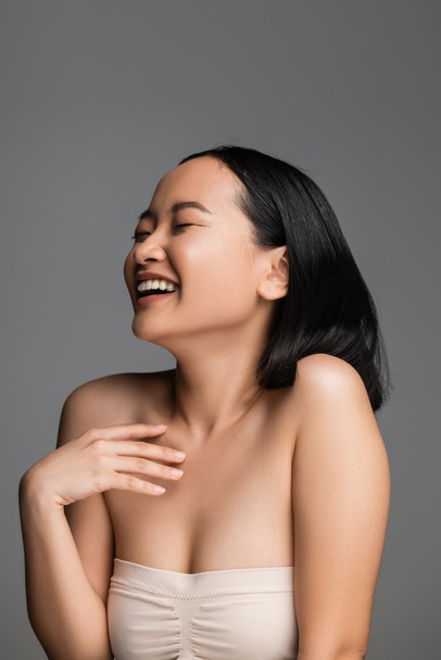 взволнованная азиатская женщина с закрытыми глазами, держась за руку рядом с грудью и смеясь изолированы на серый - Фото, изображение