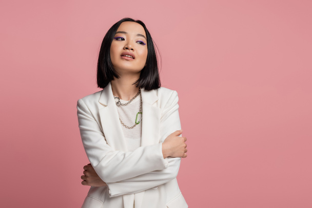 brunette asian woman in white jacket and necklaces standing with crossed arms and looking away isolated on pink - Photo, image