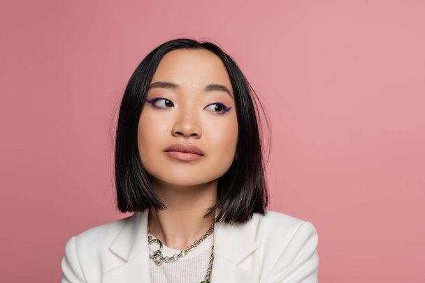 portrait of young asian woman with blue eyeliner and silver necklaces looking away isolated on pink - Photo, Image