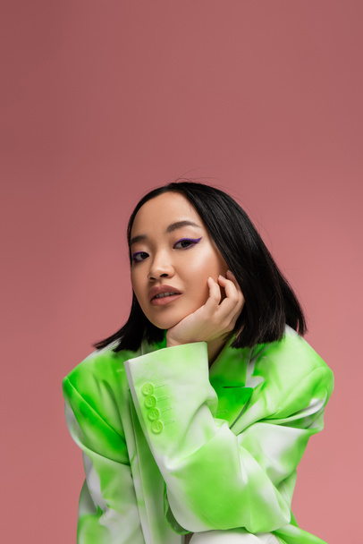 portrait of brunette asian woman in green and white blazer and makeup with blue eyeliner isolated on pink - Foto, imagen