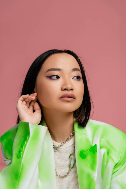 portrait of brunette asian woman in green and white jacket and makeup looking away isolated on pink - Φωτογραφία, εικόνα
