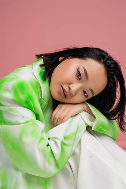 portrait of young asian woman in green and white outfit and visage with blue eyeliner looking at camera isolated on pink - Photo, image