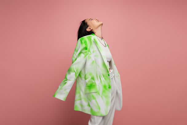 young and happy asian woman in green and white oversize blazer posing with raised head isolated on pink - Foto, Imagem