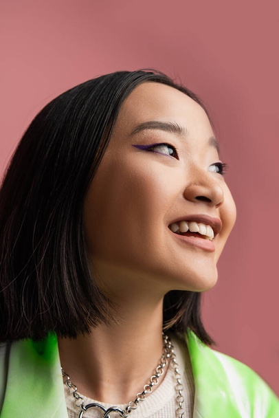 portrait of cheerful asian woman in metal necklaces and makeup looking away isolated on pink - Foto, Bild