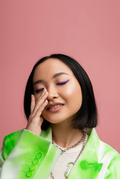 portrait of smiling asian woman with closed eyes touching perfect face with blue eyeliner isolated on pink - Zdjęcie, obraz