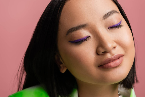 крупным планом портрет красивой азиатской женщины с идеальной кожей и синей подводкой для глаз на закрытых глаза изолированы на розовый - Фото, изображение