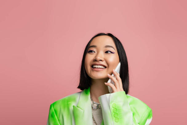 fashionable asian woman smiling and looking away while talking on mobile phone isolated on pink - Фото, зображення