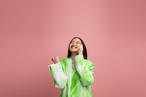взволнованная азиатская женщина в стильной белой и зеленой куртке разговаривая по мобильному телефону и показывая победу жест изолирован на розовый - Фото, изображение