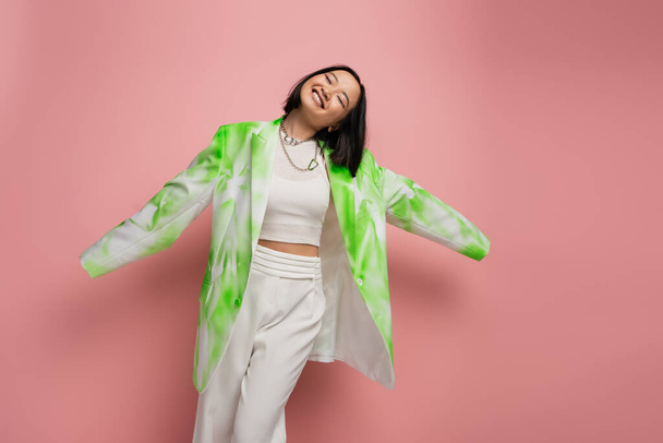 joyful asian woman in trendy oversize jacket and white pants posing with closed eyes and outstretched hands on pink background - Foto, Imagen
