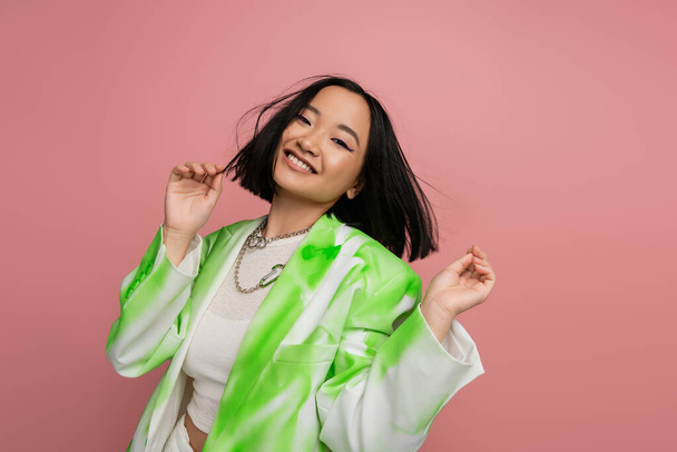 cheerful asian woman in stylish green and white jacket and silver necklaces posing isolated on pink - Photo, image