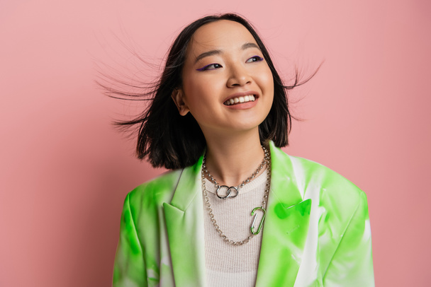 joyful asian woman in trendy outfit and makeup with blue eyeliner looking away on pink background - Foto, afbeelding