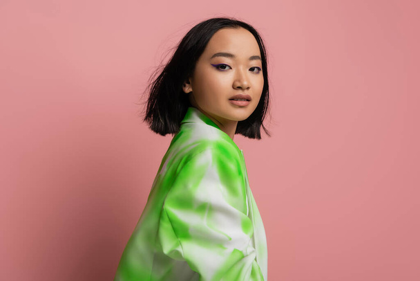 brunette asian woman in stylish green and white jacket looking at camera isolated on pink - Foto, Bild