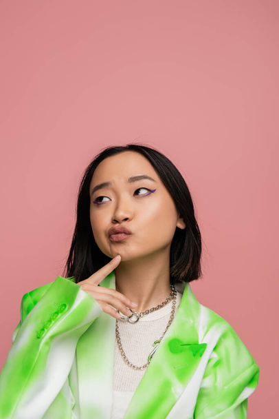 thoughtful asian woman in trendy blazer and metal necklaces touching chin and looking away isolated on pink - Foto, immagini