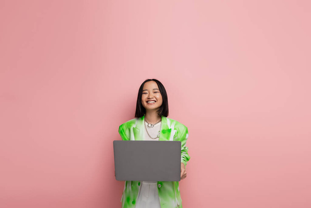 cheerful asian woman in green and white jacket smiling near laptop isolated on pink - Фото, зображення