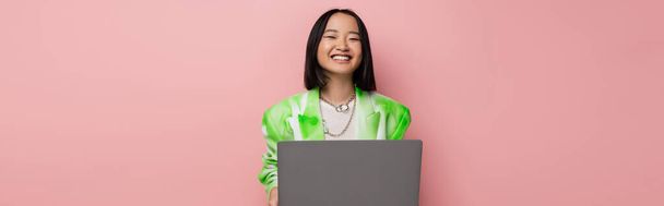緑と白のジャケットを着た明るいアジアの女性がピンクとバナーに囲まれたノートパソコンの近くで笑っています - 写真・画像