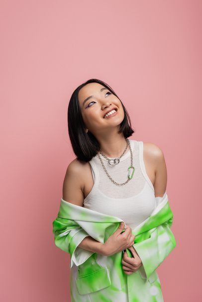happy asian woman in white tank top and metal necklaces looking away isolated on pink - Foto, immagini