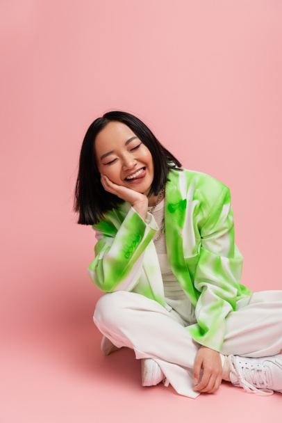 cheerful asian woman in green and white jacket sitting with crossed legs and sticking out tongue on pink background - Fotó, kép