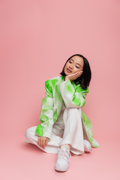 pensive asian woman in white pants and trendy jacket sitting with hand near face on pink background - Valokuva, kuva