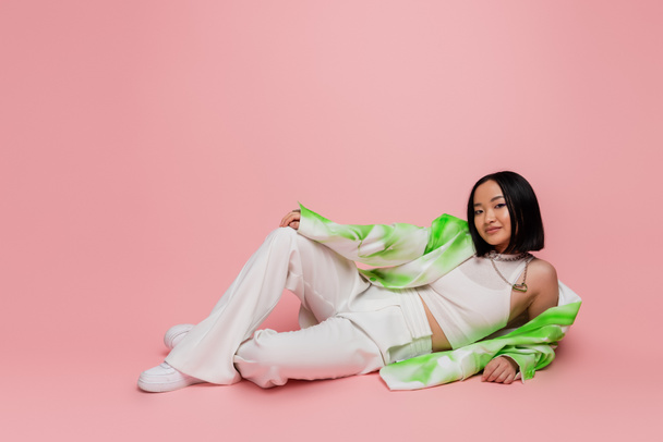 full length of smiling asian woman in white pants and fashionable jacket lying on pink background - Foto, Bild