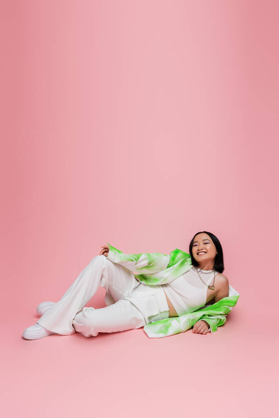 full length of asian model in white pants and trendy jacket lying and looking at camera on pink background - Photo, Image
