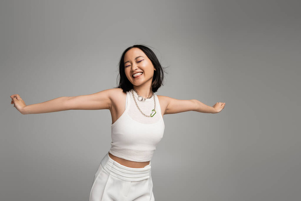 young and happy asian woman in white tank top sticking out tongue while standing with outstretched hands isolated on grey - Photo, image