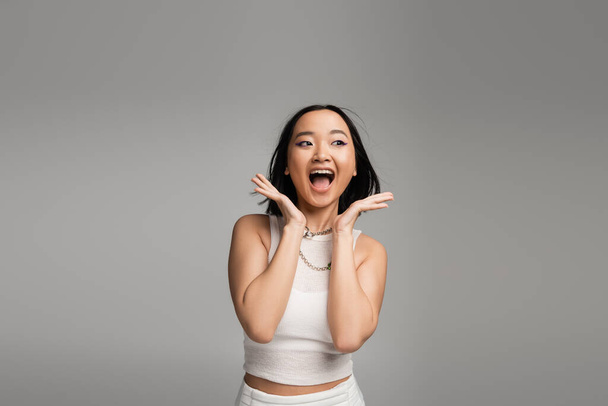 excited asian woman in white tank top holding hands near face and screaming isolated on grey - Foto, imagen