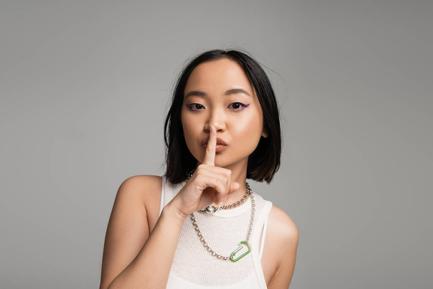 young asian woman in white tank top and metal necklaces showing hush sign isolated on grey - Photo, image