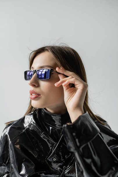 молодая женщина в черной латексной куртке регулирует модные солнцезащитные очки, изолированные на сером - Фото, изображение