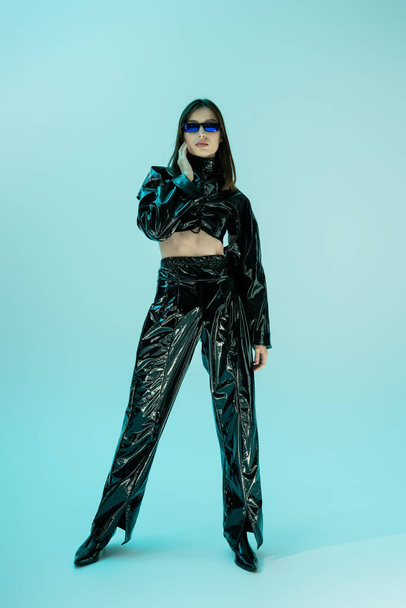 full length of stylish young woman in black latex clothing and sunglasses standing on blue  - Φωτογραφία, εικόνα