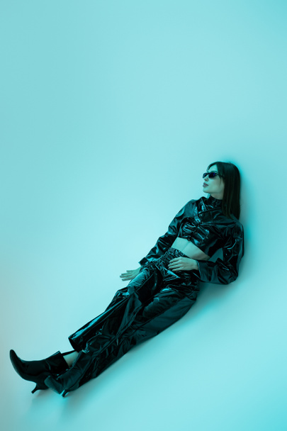 full length of stylish young woman in black latex clothing and high heels sitting on blue  background  - Fotó, kép