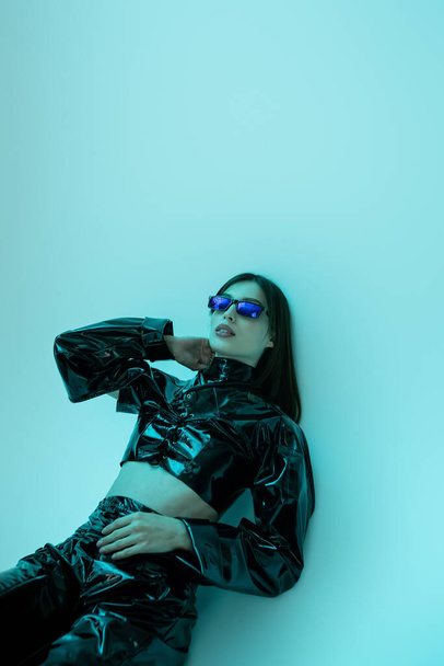 stylish young woman in sunglasses and black latex clothing sitting on blue background  - Фото, изображение