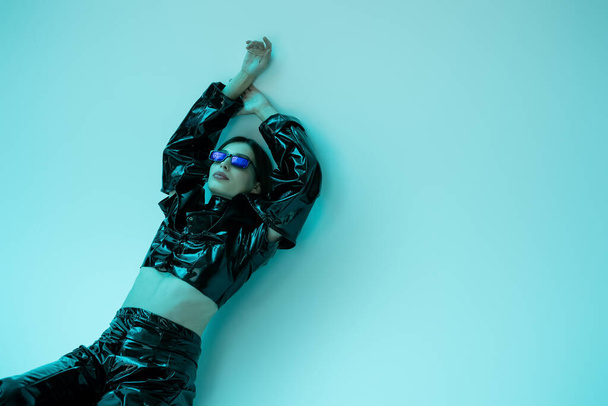 stylish young woman in sunglasses and black latex clothing posing with raised hands on blue background  - Valokuva, kuva
