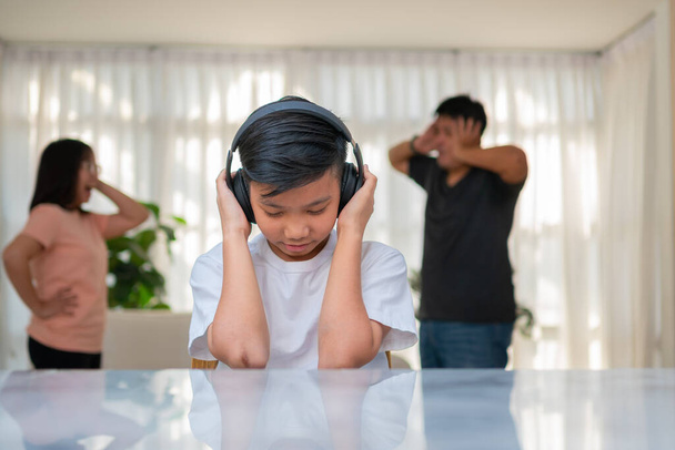 Aziatisch jongetje draagt een koptelefoon en speelt luide muziek. om thuis geen ruzie te horen terwijl ouders ruzie hebben of ruzie hebben. Ongelukkig probleem in het gezin, Huiselijke problemen in het gezin. - Foto, afbeelding