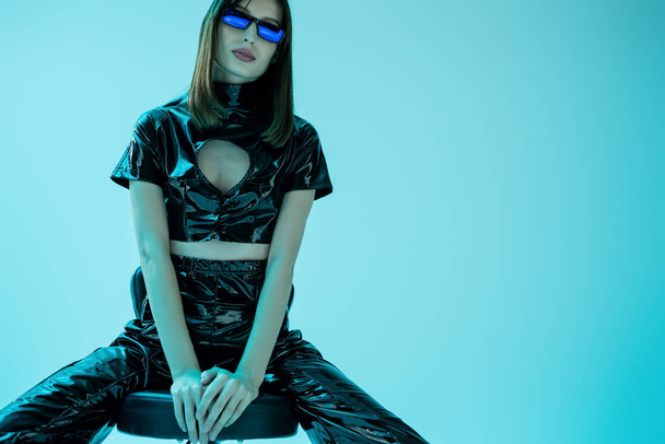 stylish young woman in latex crop top and trousers sitting on stool while looking at camera isolated on blue - Foto, immagini