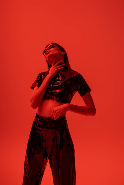 молодая и стильная женщина в топе из латекса и брюках, касающихся шеи, изолированных на красном - Фото, изображение